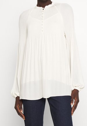 Valkoinen pitkähihainen pusero, jossa on laskostettu yläosa, napitettu edestä ja väljä malli, käytetty tumma sinisten farkkujen kanssa.