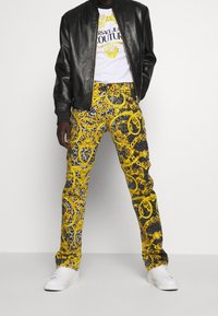 Svart läderbomberjacka, vit grafisk T-shirt, gula mönstrade byxor med guldkedjemönster och vita sneakers.