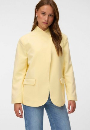 Frau mit langen braunen Haaren trägt einen hellgelben Blazer mit zwei Fronttaschen und blauen Jeans vor einfachem Hintergrund.