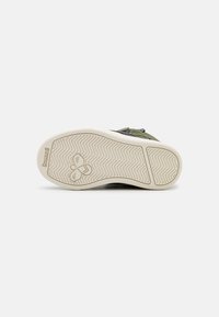 Suela de goma de una Zapatilla verde que muestra un patrón de huellas en zigzag, logo de abeja y la marca "hummel" sobre un fondo blanco.