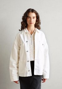 Weißer Baumwolljacke mit vorderen Taschen und Druckknöpfen, über einem gestreiften Oberteil getragen, kombiniert mit einem dunklen Rock. Strukturierter Stoff.