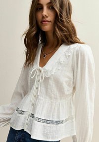 Jonge vrouw in een witte, gestructureerde blouse met kantafwerking, gerimpelde kraag, parelknopen en een hartvormige hangerketting.