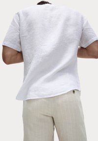 Camisa corta de lino blanco con un ajuste relajado, que presenta una sutil textura y un dobladillo curvado, combinada con pantalones de rayas finas en beige claro.