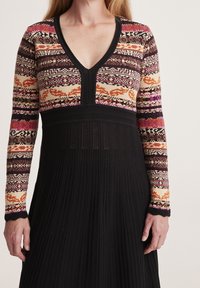 Květinové šaty s dlouhým rukávem a pletenou horní částí v červené, oranžové a fialové barvě. Černá žebrovaná sukně vyniká jemnou vertikální strukturou. Design s výstřihem do V.