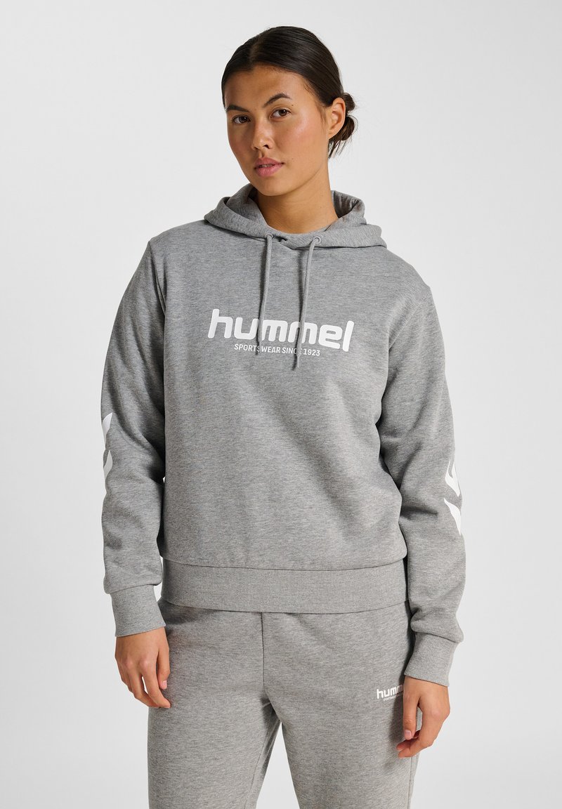 Vrouw in grijze hummel sportoutfit met een hoodie en bijpassende sweatpants, staande tegen een effen witte achtergrond.