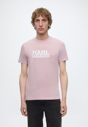 Mladý muž se světle hnědými vlasy, který má na sobě světle růžové tričko s logem "KARL LAGERFELD" a černé džíny, stojí před bílým pozadím.
