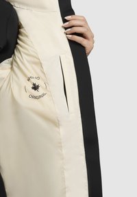 Veste noire avec intérieur crème, dotée d'une poche latérale, d'un logo de marque et d'une texture lisse. L'extérieur présente un design épuré et aérodynamique.