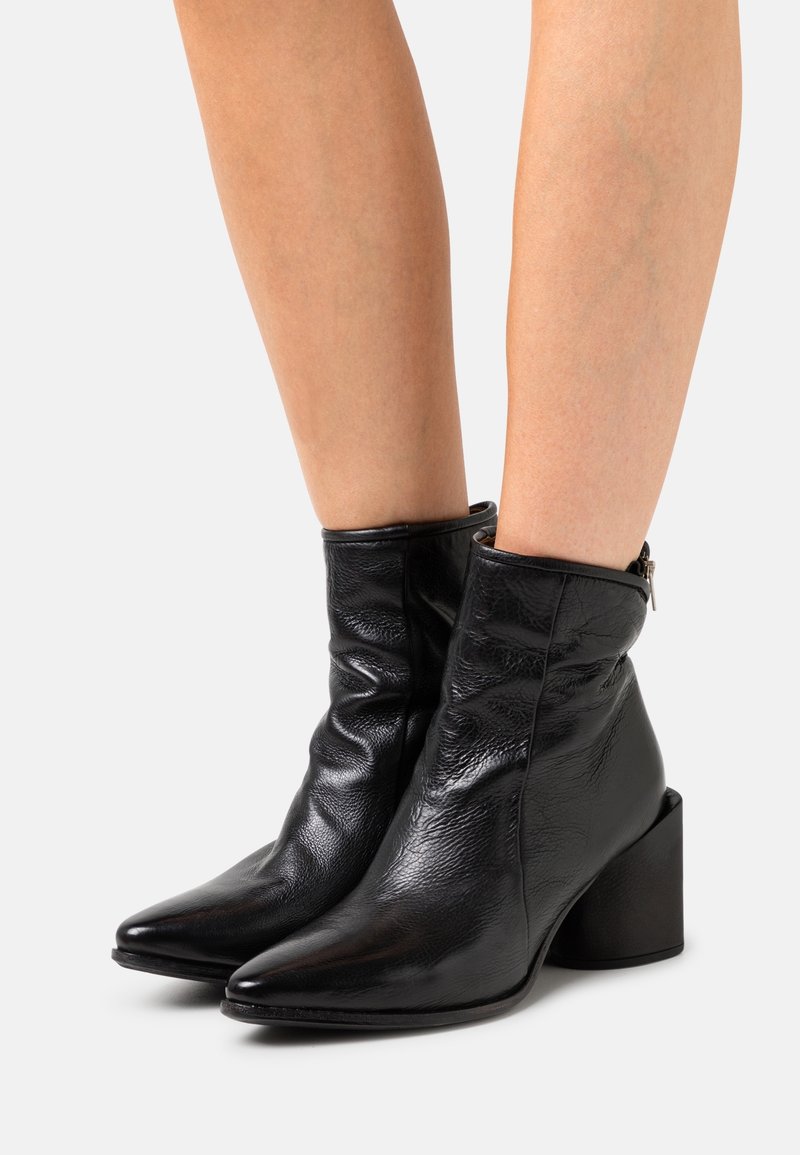 Bottes noires en cuir jusqu'à la cheville, avec des bouts pointus, des talons carrés et des fermetures éclair à l'arrière, portées sur des jambes nues sur un fond blanc.