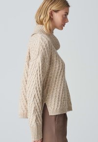 Beige Kabelstrickpullover mit hohem Kragen, rippgestrickten Bündchen und Saum sowie einem leicht verkürzten Schnitt. Weiche Textur mit einem gemütlichen Erscheinungsbild.