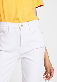 Jeans de denim blanco con cierre de botón frontal y cinco bolsillos. Textura suave y diseño ajustado, combinados con una camisa amarilla brillante.