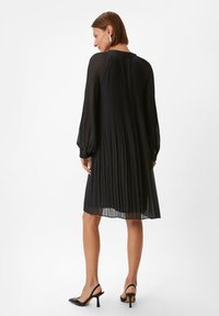 Schwarzes, plissiertes Kleid mit langen Ärmeln, leichtem Material, rundem Ausschnitt und knöchellangem Saum, kombiniert mit schwarzen, hochhackigen Sandalen.
