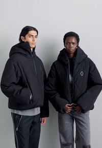 Vestes matelassées noires avec capuches, présentant des accents en forme de diamant et un design matelassé. Portées par-dessus des tenues décontractées avec différents styles de pantalons.