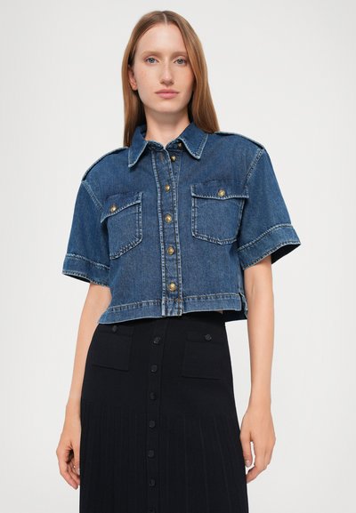 Kuttet denimskjorte med korte ermer, blå farge, gullknapper, to frontlommer og klassisk krage; kombinert med et svart skjørt.