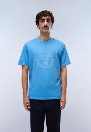 Mann mit lockigem Haar und Schnurrbart trägt hellblaues Grafik-T-Shirt und dunkle Hose, steht vor schlichtem hellgrauem Hintergrund.