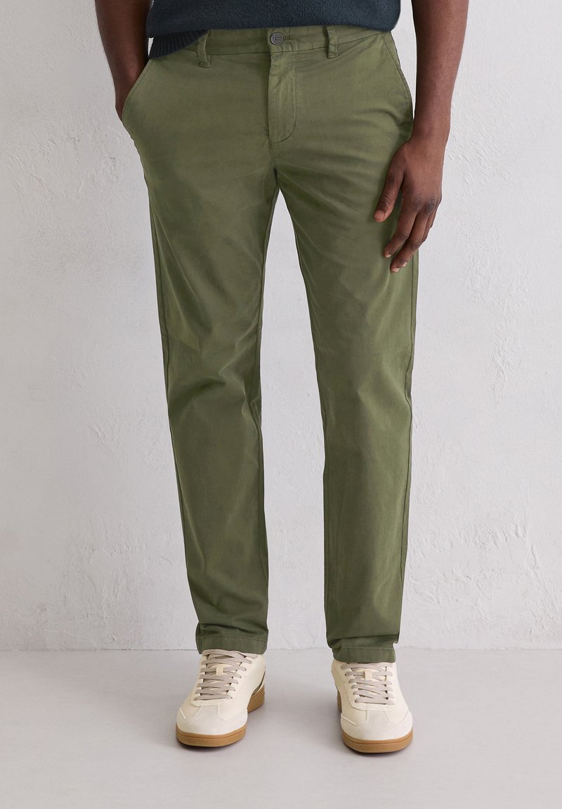 Zielone bawełniane spodnie chino z bocznymi kieszeniami, o wąskim kroju, z płaskim przodem, szlufkami na pasek i gładką fakturą, w połączeniu z beżowymi tenisówkami.