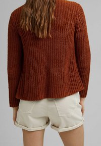 Rust-colored knitted cardigan with a textured finish; features long sleeves and a flared hem, paired with rolled beige shorts.