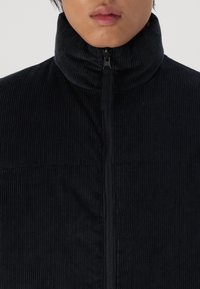 Chaqueta de pana negra con un alto collar, cierre de media cremallera en la parte delantera y costillas verticales texturizadas en la superficie de la tela.