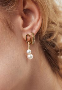 Złota kolczyk w kształcie obręczy charakteryzuje się teksturowanym paskiem oraz zwisającym łańcuszkiem z dwiema białymi perłami, ukazując mix materiałów i elegancki design.