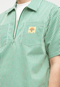 Πράσινο και λευκό πουκάμισο με κάθετες ρίγες, κοντά μανίκια, φερμουάρ μπροστά και τσέπες στο στήθος, φορεμένο πάνω από ένα λευκό μπλουζάκι με πολύχρωμο σχέδιο.