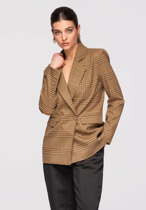 Kvinde iført brun ternet blazer og sorte bukser poserer med den ene hånd nær kraven og den anden i lommen mod en neutral baggrund.