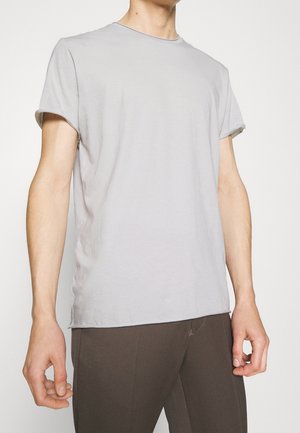 Person iført en lysegrå kortærmet t-shirt og mørkebrune bukser, stående med armene afslappet ned langs siden.