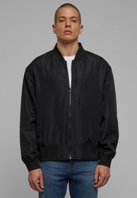 Veste bomber noire en tissu lisse, dotée d'une fermeture éclair à l'avant, de poignets et d'une taille côtelés. Deux poches latérales et un col classique.