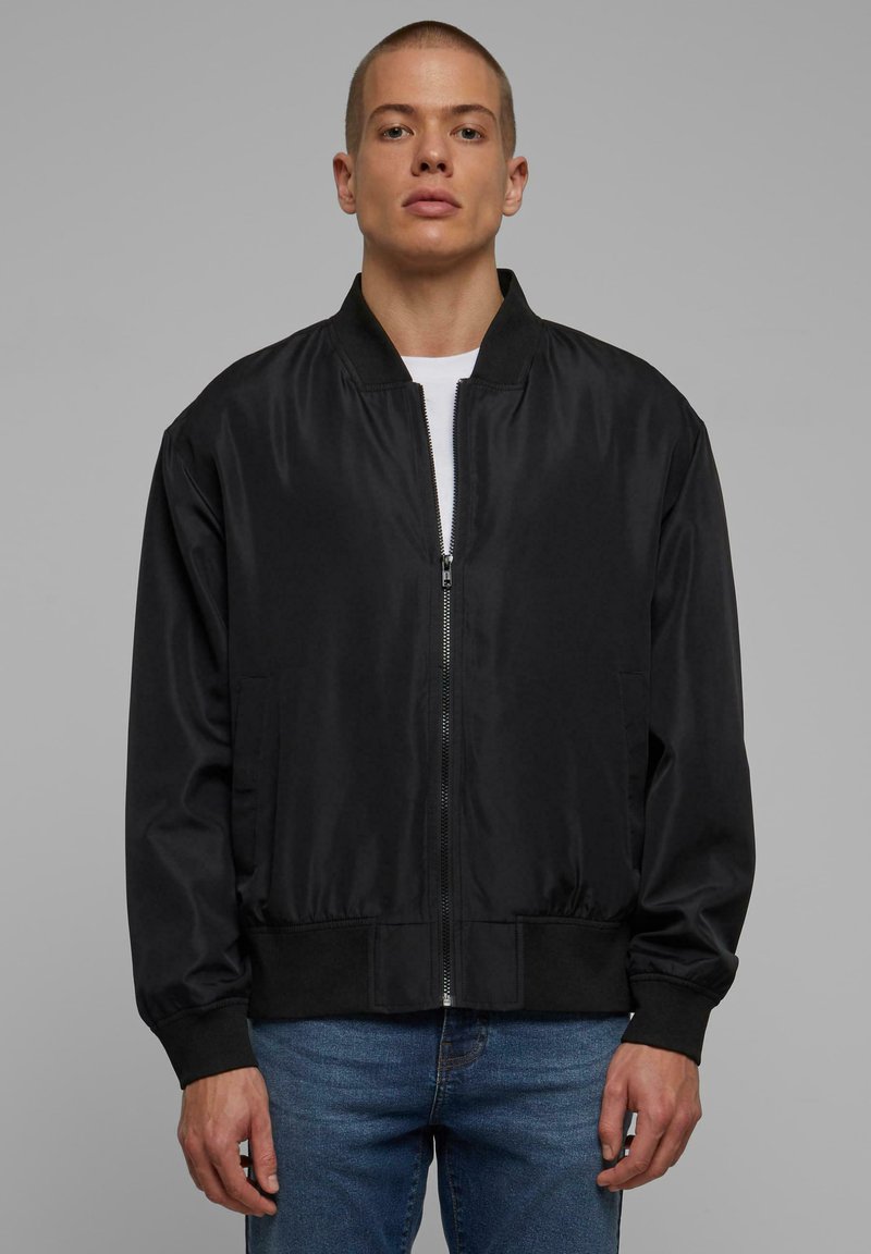 Veste bomber noire en tissu lisse, dotée d'une fermeture éclair à l'avant, de poignets et d'une taille côtelés. Deux poches latérales et un col classique.