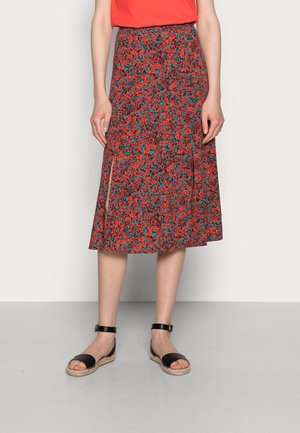 Femme portant une jupe midi rouge à motifs floraux avec des fentes latérales, des sandales noires à bride cheville et un haut rouge, debout contre un mur uni.
