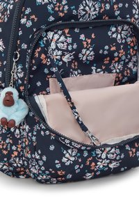 Mochila de fondo marino con patrón floral, con un bolsillo con cremallera, forro rosa y un llavero de mono azul. Presenta una tela texturizada y acentos en contraste.