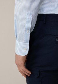 Lichtblauwe en witte gestreepte shirtmouw met een knoopsluiting, gecombineerd met donkerblauwe broek, die de soepele stoftextuur benadrukt.