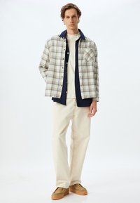 Camisa de cuadros en beige y azul marino con un cierre de botones, combinada con jeans color blanco roto y zapatos marrones. Presenta un bolsillo en el pecho y un corte relajado.