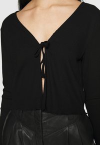 Schwarzes, geripptes Langarmoberteil mit tiefem V-Ausschnitt und vorderer Knoten-Detail. Der Stoff ist weich und der Saum leicht ausgefranst für einen lässigen Look.