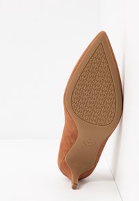 Chaussure à talon kitten en daim marron à bout pointu reposant sur le côté, montrant la semelle avec un motif répété du logo "MK".
