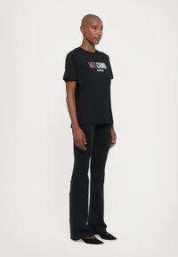 Juodas medvilnės T-marškinėliai trumpomis rankovėmis, su daugiakomponiu "Moschino Jeans" logotipu priekyje, derinami su juodomis prigludusiomis kelnėmis ir bateliais.
