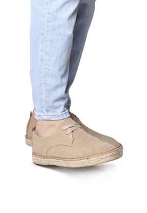 Beige espadrille-style shoes made of textured canvas with a round toe, lace-up front, and woven jute sole, set against light denim.