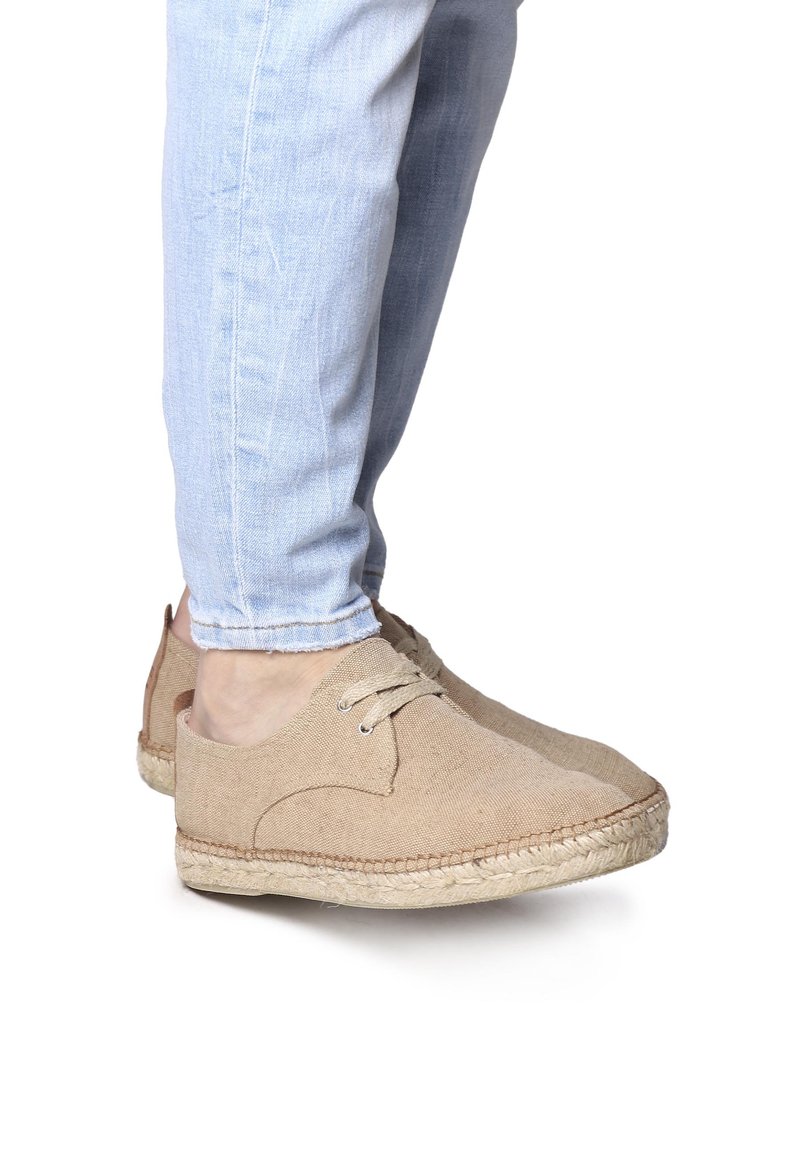 Scarpe beige in stile espadrilla realizzate in tela texture, con punta rotonda, parte anteriore allacciata e suola in juta intrecciata, su uno sfondo di denim chiaro.