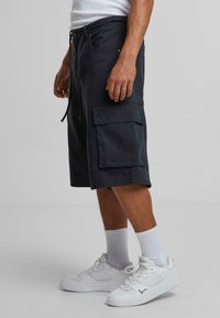 Dark gray cargo shorts made of cotton, featuring a relaxed fit, side pockets, drawstring closure, and knee-length cut. Paired with white sneakers.