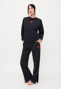 Vrouw staat blootvoets in een zwarte lange mouwtop en broek met rode Hugo-logo patches op de borst en dij.