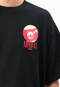 Sort bomulds t-shirt med et rundt grafik i rød, gul og hvid med Adidas-logoet og teksten "ADIDAS ORIGINALS." Korte ærmer.