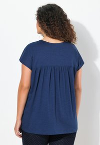 Tmavě modrý top s krátkým rukávem a nařaseným designem vzadu. Měkká texturovaná látka s volným střihem, v kombinaci s kalhotami s puntíky.