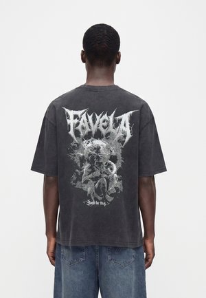 Person trägt ein dunkles, übergroßes T-Shirt mit weißem grafischem Text "Favela" und einer künstlerischen Skizze eines zusammengerollten Fötus auf dem Rücken.