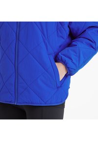 Person wearing a blue quilted jacket with a zipper, hand partially inside the side pocket, and black pants visible below.