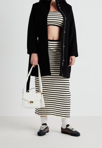 Černý dlouhý kabát přes černobíle pruhovaný crop top a sukni. Bílá kabelka se zlatými akcenty. Tmavé boty na šněrování a bílé ponožky.