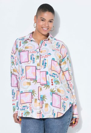 Smilende person med kort hår iført øreringe med ringe, en farverig skjorte med knapper med tropiske og vintage bilmotiver samt blå jeans.