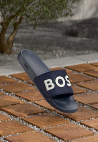 Tengerészkék csúszó-szandál, texturált pánttal, amelyen fehér "BOSS" felirat található, gumiból készült, sima talpbetéttel és lapos talppal.