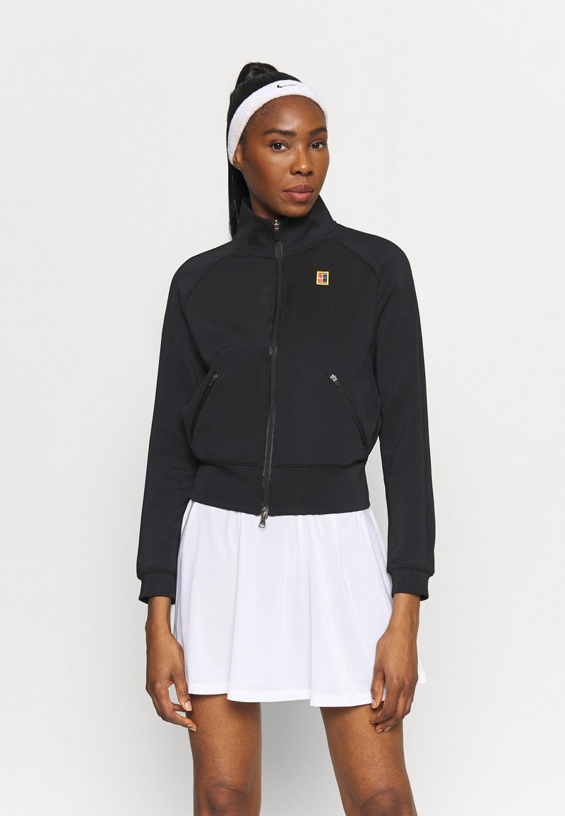 Schwarze Zip-Jacke mit figurnahem Schnitt, gerippten Bündchen und einem kleinen Logopatch. Tragen sie über einem weißen, ausgestellten Rock, was einen sportlichen Look zeigt.