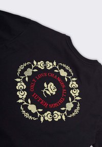 Tessuto nero con una ghirlanda floreale ricamata in crema e testo rosso al suo interno che recita "SOLTANTO L'AMORE CAMBIA TUTTE LE COSE IUTER," circondando un design centrale di rosa.