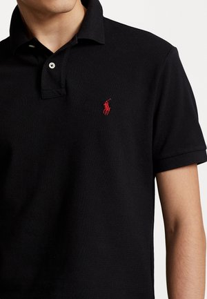 Mężczyzna noszący czarną koszulkę polo z krótkim rękawem, z czerwonym wyszywanym logo na lewej piersi i dwoma białymi guzikami na kołnierzyku.