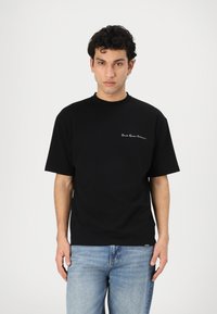 Black cotton t-shirt with a crew neckline and short sleeves, featuring white text embroidered on the left chest area. Paired with light denim jeans.