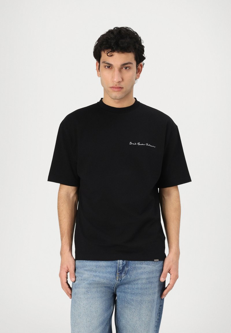 Black cotton t-shirt with a crew neckline and short sleeves, featuring white text embroidered on the left chest area. Paired with light denim jeans.